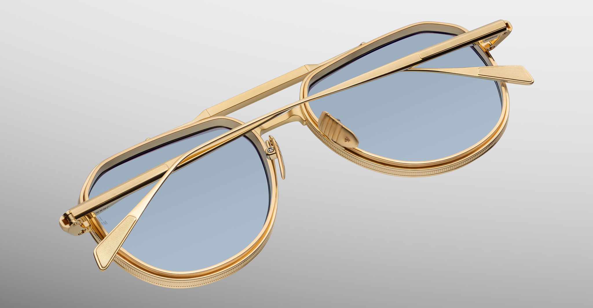 The Jacques Marie Mage Commander sunglasses, featuring gold frames and blue-tinted lenses, are displayed upside down with folded arms against a smooth gradient background from light gray to white.