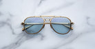 A pair of Jacques Marie Mage Commander sunglasses with gold frames and light blue lenses rests on a white marble surface, reflecting the marble pattern in the lenses.