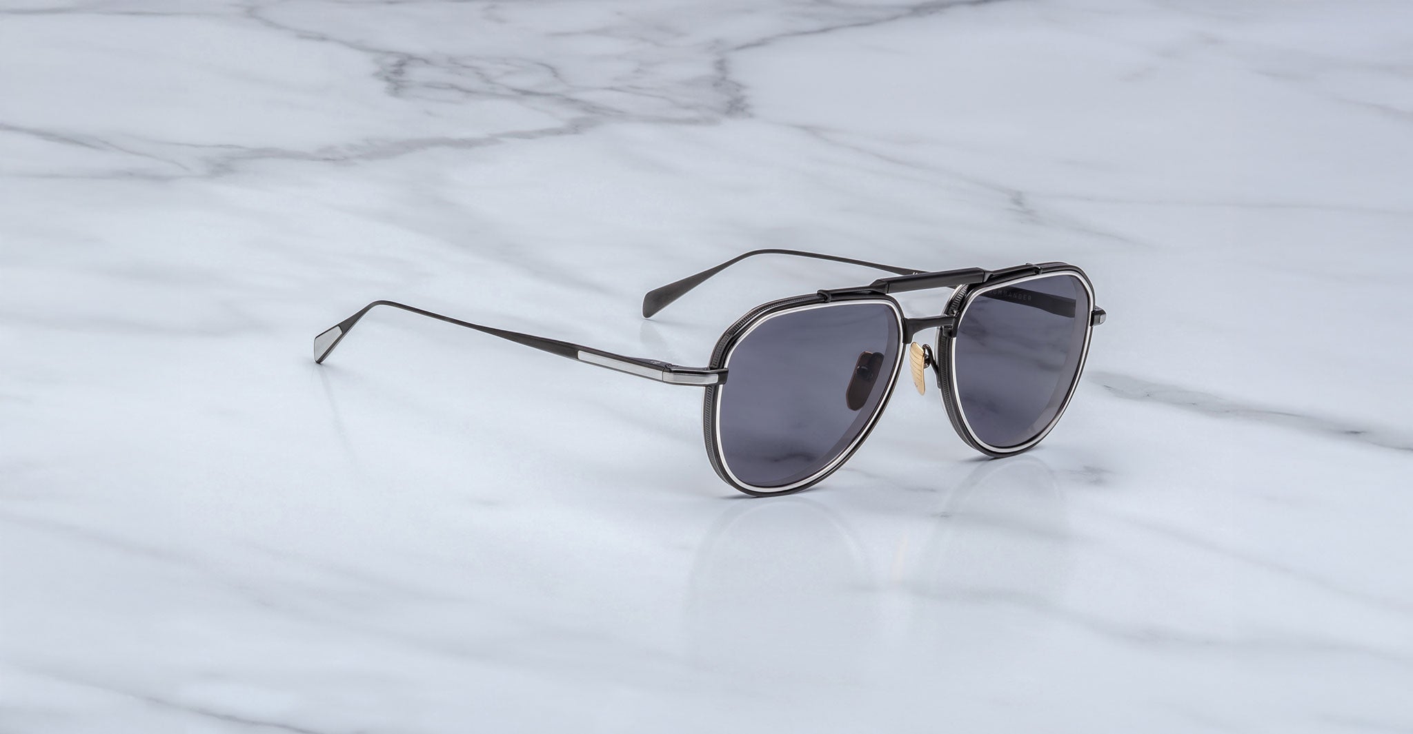 The Jacques Marie Mage Commander, featuring black aviator frames and dark lenses, rests on a white marble surface with gray veins.