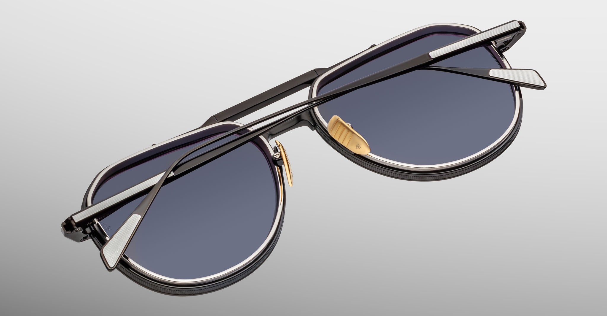A pair of Jacques Marie Mage Commander sunglasses with round dark lenses and black-silver frames, shown folded with the crossed arms over the lenses, set against a light gradient background.