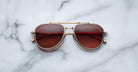 A pair of Jacques Marie Mage Commander gold-framed aviator sunglasses with red-tinted lenses rests on a white marble surface.