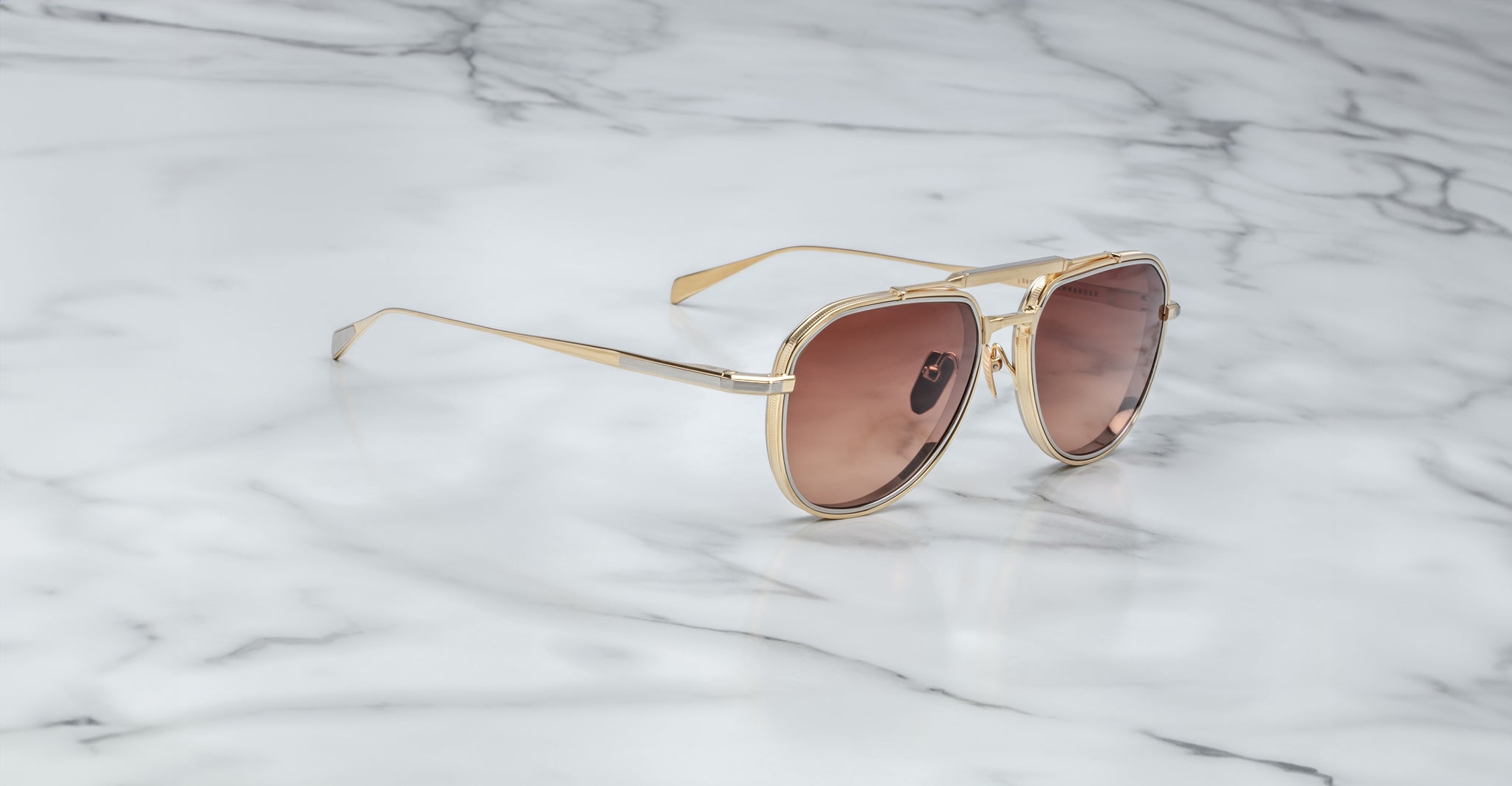 A Jacques Marie Mage Commander gold-framed sunglasses with brown-tinted lenses rest on a white marble surface with gray veining.