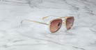 A Jacques Marie Mage Commander gold-framed sunglasses with brown-tinted lenses rest on a white marble surface with gray veining.