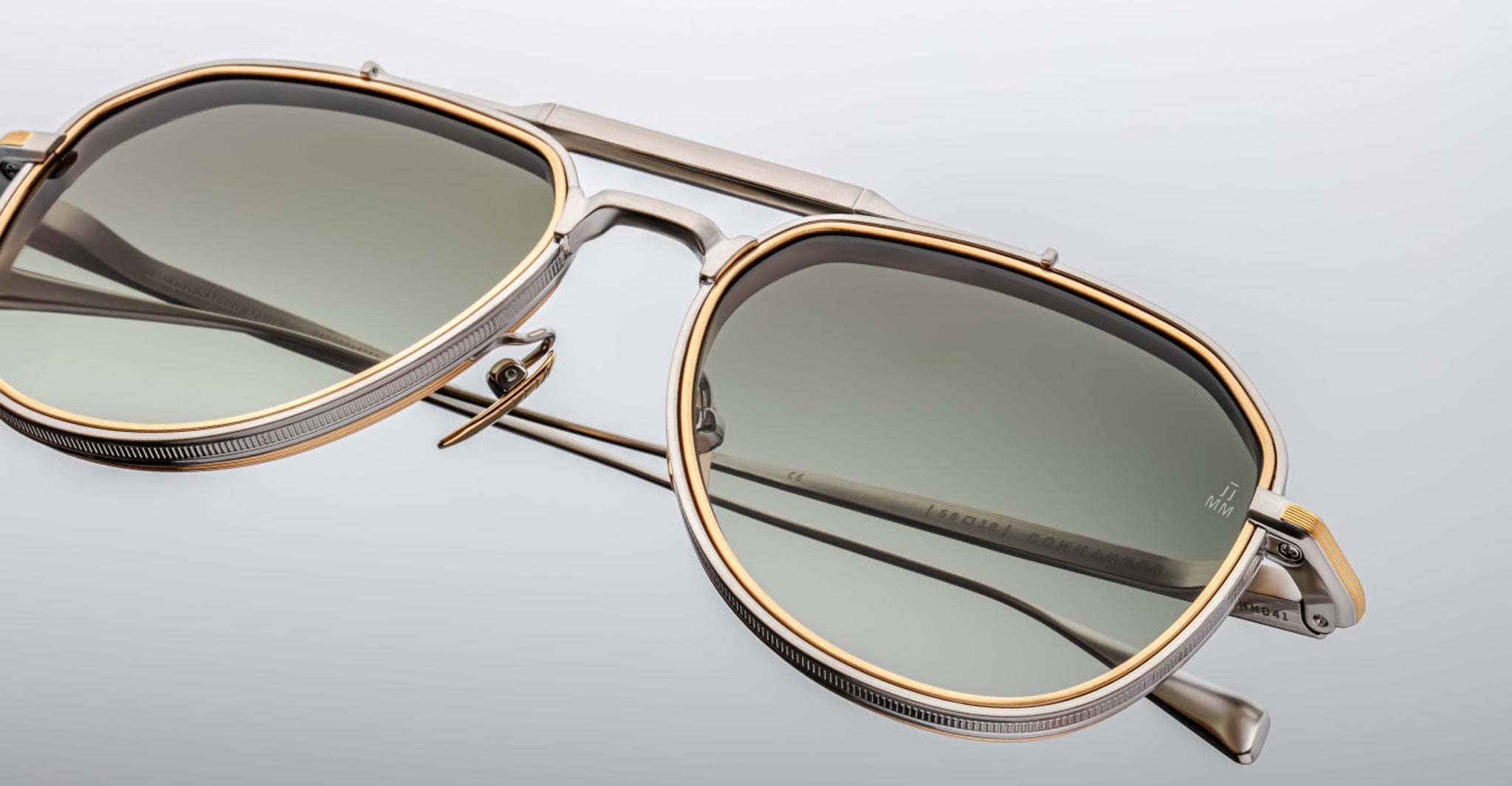 Close-up of Jacques Marie Mage Commander sunglasses, showcasing a metallic silver and gold frame with green-tinted lenses and intricate bridge and temple details, set against a plain light gray background.