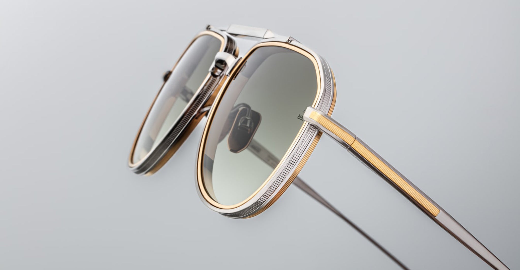 A close-up of the Jacques Marie Mage Commander sunglasses, featuring metal and gold-toned frames with green-tinted lenses, shown from an angle that highlights the temples and side details against a plain background.