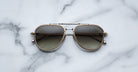 A pair of Jacques Marie Mage Commander aviator sunglasses with metal frames and tinted lenses rests on a white marble surface with gray veining.