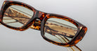 Close-up of Jacques Marie Mage Corniche rectangular tortoiseshell eyeglasses with light-colored lenses, reflecting gold metallic details in the background, set on a plain light gray surface.