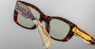 A close-up of Jacques Marie Mage Corniche tortoiseshell sunglasses with rectangular green-tinted lenses and gold metal temple detailing, set against a plain light gray background.
