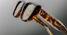 Close-up of Jacques Marie Mage Corniche eyeglasses showing the brown tortoiseshell pattern on the front and side. Set against a plain gray background, the image highlights the frames detailed texture and craftsmanship.