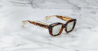 A pair of Jacques Marie Mage Corniche rectangular tortoiseshell sunglasses with thick frames and green-tinted lenses rests on a white marble surface.