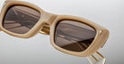Close-up of Jacques Marie Mage Corniche sunglasses—beige, rectangular frames with thick rims and dark brown lenses reflecting geometric patterns, featuring metallic hinge details on the temples against a neutral background.
