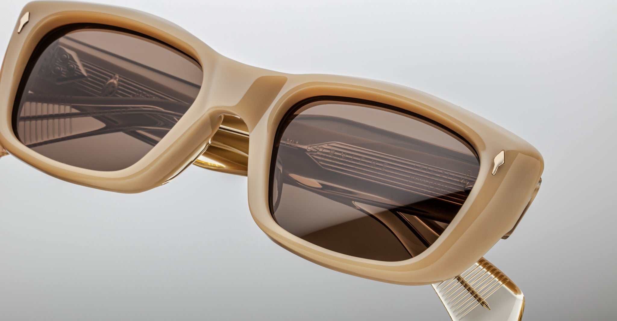 Close-up of Jacques Marie Mage Corniche sunglasses—beige, rectangular frames with thick rims and dark brown lenses reflecting geometric patterns, featuring metallic hinge details on the temples against a neutral background.