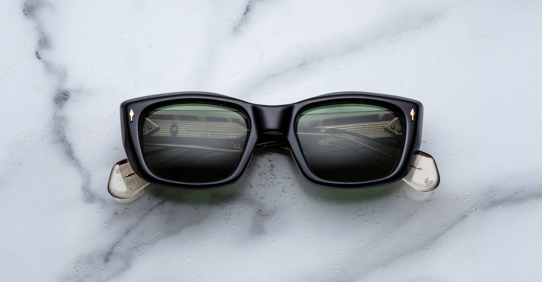 The Jacques Marie Mage Corniche sunglasses—a black frame with green-tinted lenses and transparent arm tips—rest on a white marble surface, their lenses reflecting a geometric pattern.