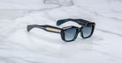 The Jacques Marie Mage Corniche sunglasses, with thick black frames, dark lenses, and metallic gold-accented temples featuring embossed branding near the hinges, rest on a white marble surface.
