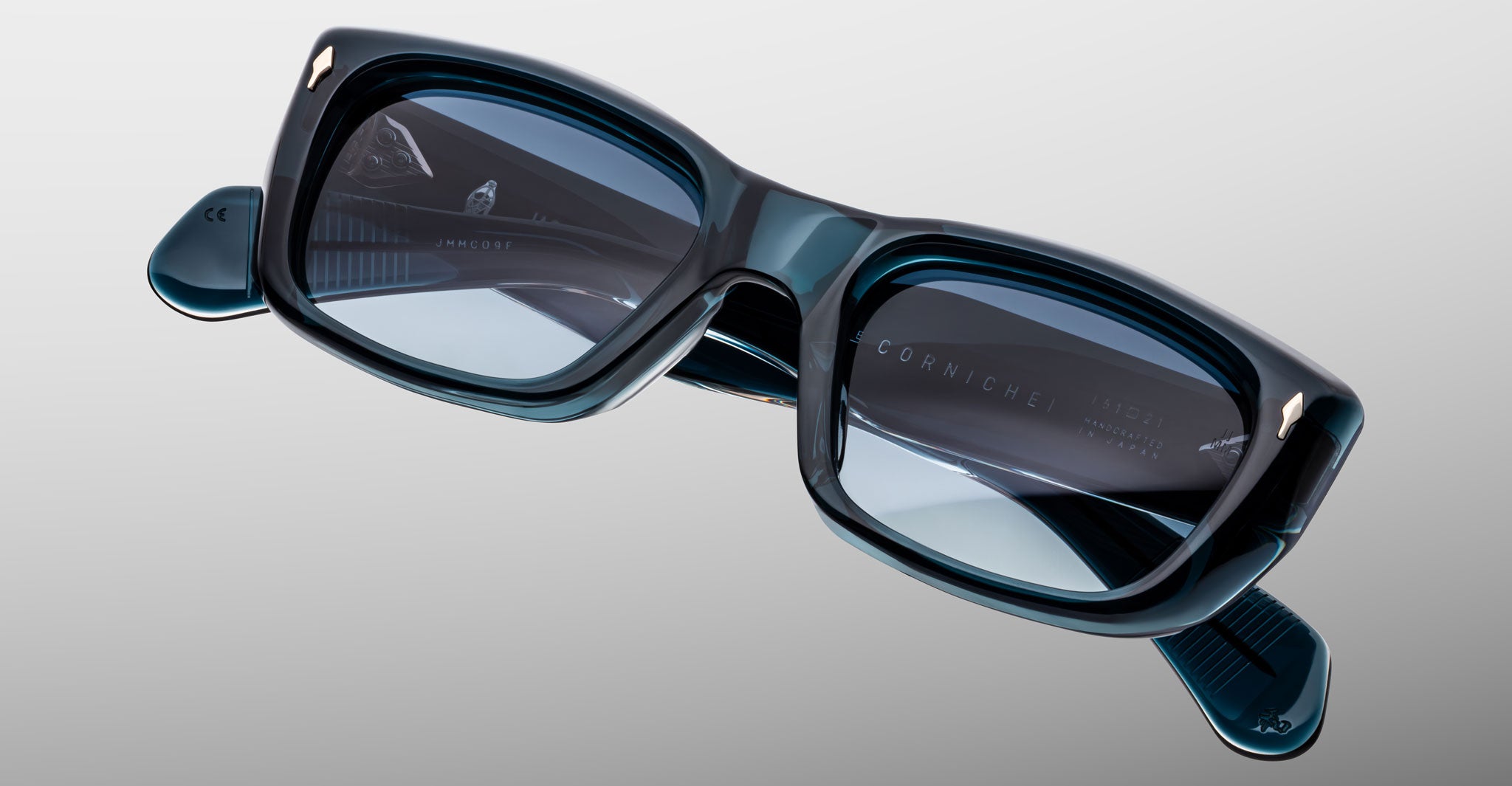 Close-up of Jacques Marie Mage Corniche rectangular, thick-framed dark teal eyeglasses with lightly tinted lenses on a light gray background. The Corniche branding is visible on the inside of the right temple.