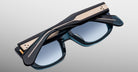 The Jacques Marie Mage Corniche sunglasses feature black frames, blue-tinted lenses, and gold metallic temple accents. Shown folded with arms on top, theyre displayed against a light grey background.