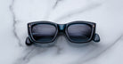 The Jacques Marie Mage Corniche sunglasses, featuring a black rectangular frame and dark lenses, rest on a white marble surface with gray veins. The arms of the sunglasses are folded.