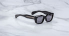 A pair of Jacques Marie Mage Corniche black rectangular sunglasses with thick frames and dark lenses rests on a white marble surface. The arms feature metallic accents with horizontal lines near the hinges.