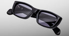 The Jacques Marie Mage Corniche sunglasses, featuring black thick-rimmed rectangular frames with dark lenses and folded arms, are displayed on a light gray background. Brand markings appear on the inside of the frames.