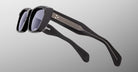 The Jacques Marie Mage Corniche sunglasses feature thick black frames and dark lenses, shown from a side and tilted angle on a plain gray background. The temples display metallic accent stripes and textured grips.