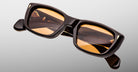 The Jacques Marie Mage Corniche sunglasses feature thick black rectangular frames and amber-tinted lenses, shown against a light grey background. CORNICHE is labeled on the inner arms along with additional small text inside.