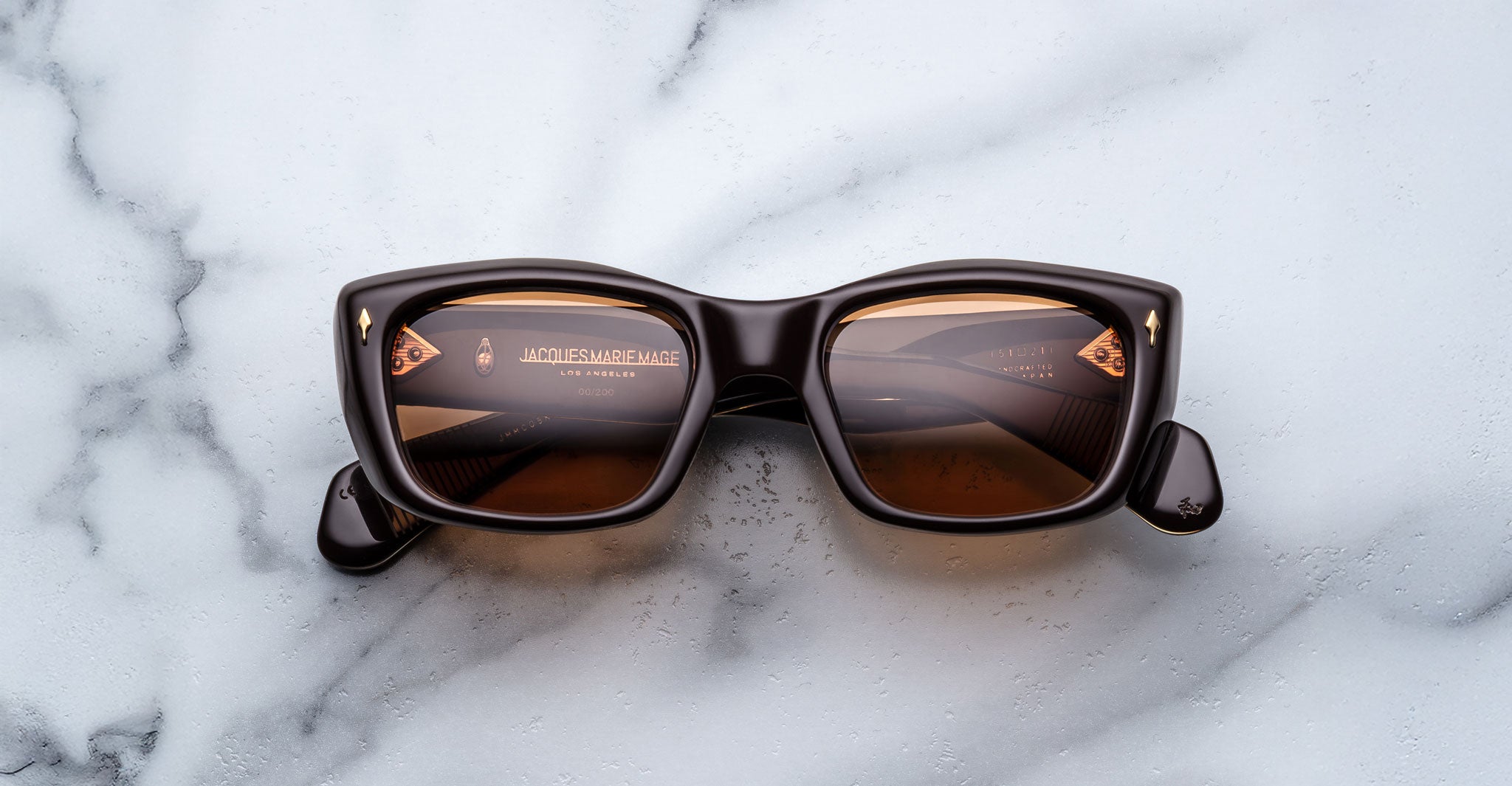 A pair of Jacques Marie Mage Corniche rectangular brown sunglasses with tinted lenses rests on a white marble surface, brand text and logo visible through the inner frames.