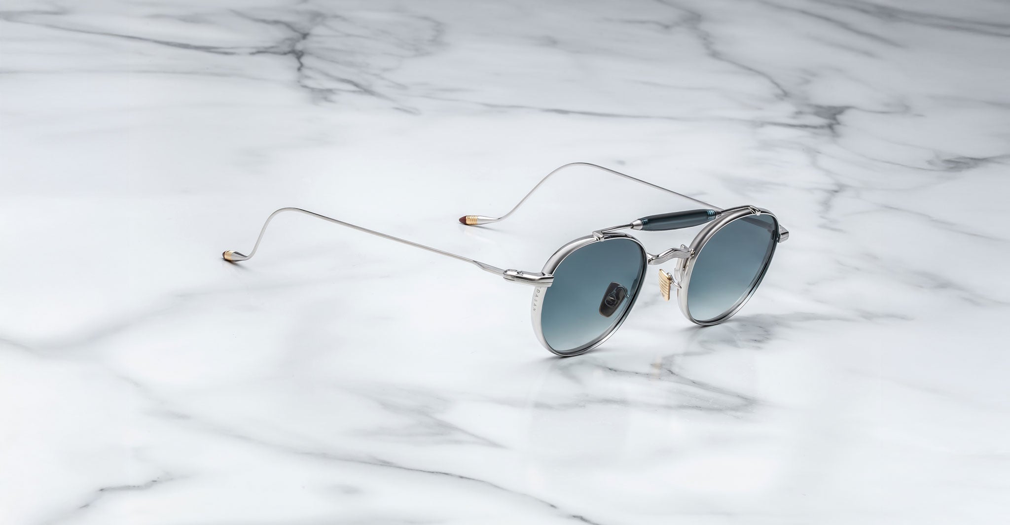 Profile view of the Jacques Marie Mage Dasan titanium sunglass in silver with blue gradient lenses on a white marbled with grey veining background
