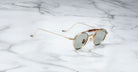 Profile view of the Jacques Marie Mage Dasan titanium sunglass in gold color with light grey lenses on a white marbled with grey veining background