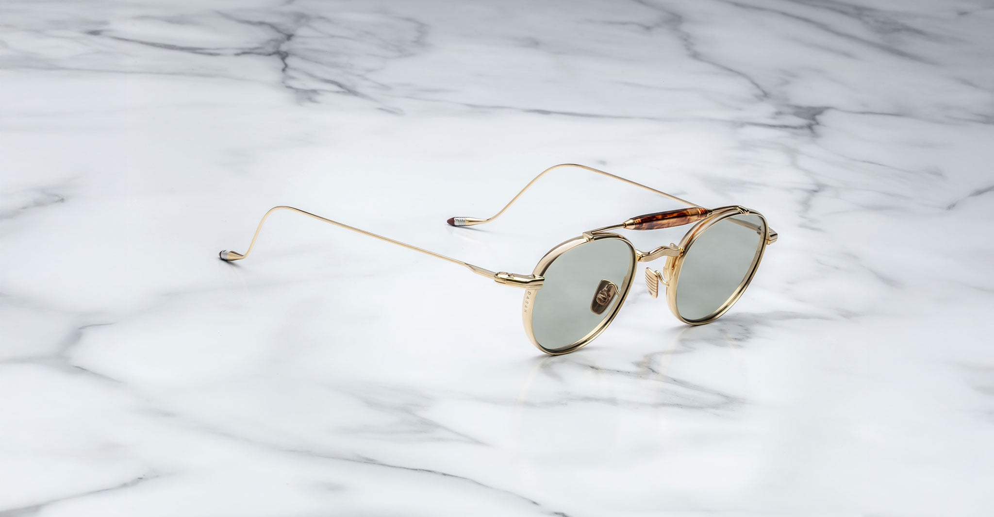 Profile view of the Jacques Marie Mage Dasan titanium sunglass in gold color with light grey lenses on a white marbled with grey veining background