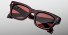 The Jacques Marie Mage Dealan 53 sunglasses, featuring square-shaped thick dark burgundy frames and reddish tinted lenses, are displayed against a light gray background.