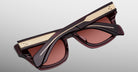 A pair of Jacques Marie Mage Dealan 53 sunglasses with thick frames, dark reddish lenses, and gold temple detailing, displayed folded on a light gray background.