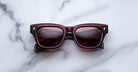 The Jacques Marie Mage Dealan 53 sunglasses, featuring a rectangular burgundy frame and dark lenses, rest on a white marble surface. Gold text and subtle decorative details accent the inside of the arms.