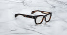 A Jacques Marie Mage Dealan 53 rectangular eyeglasses with a tortoiseshell frame rest on a white marble surface.
