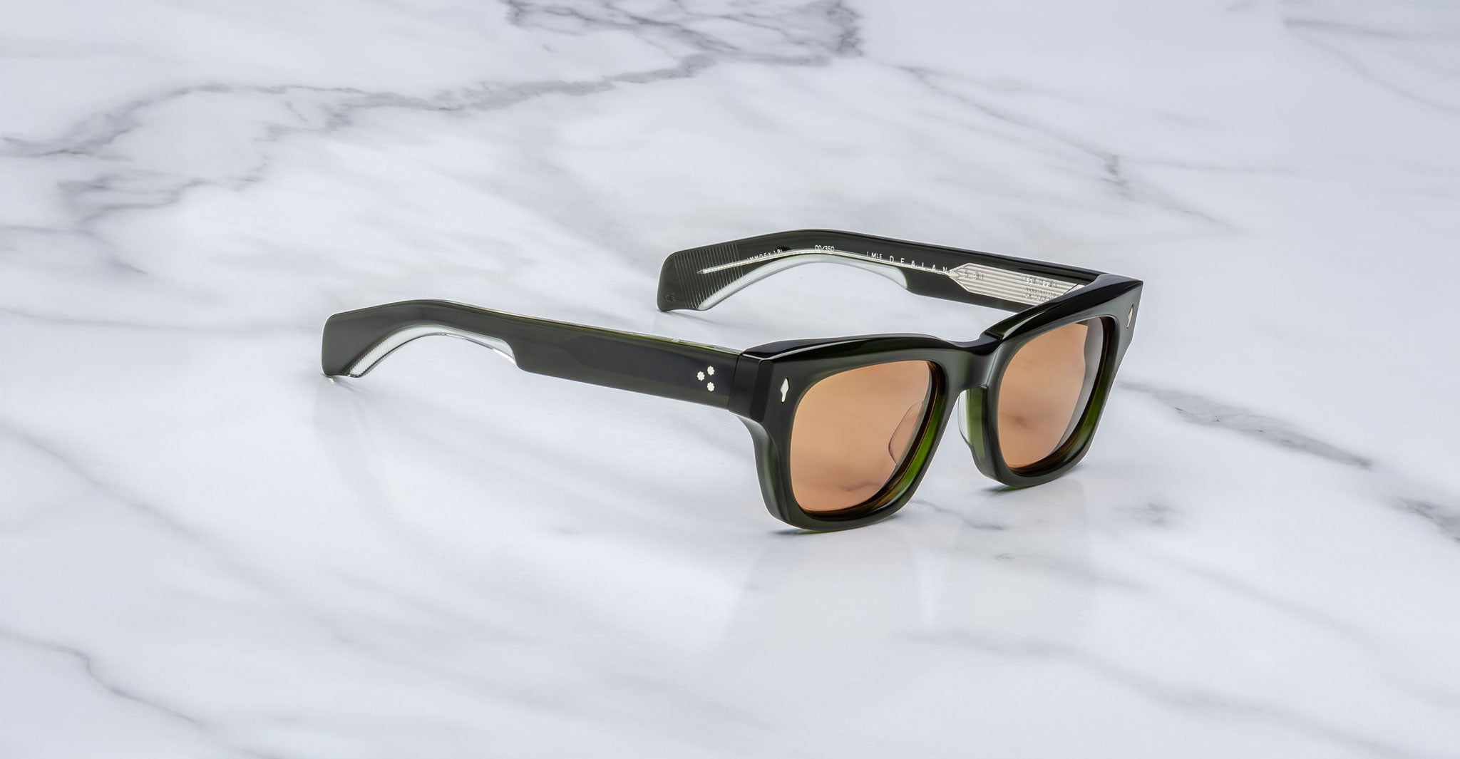 The Jacques Marie Mage Dealan 53 sunglasses, featuring chunky rectangular dark green frames and light brown tinted lenses, rest on a white marble surface.