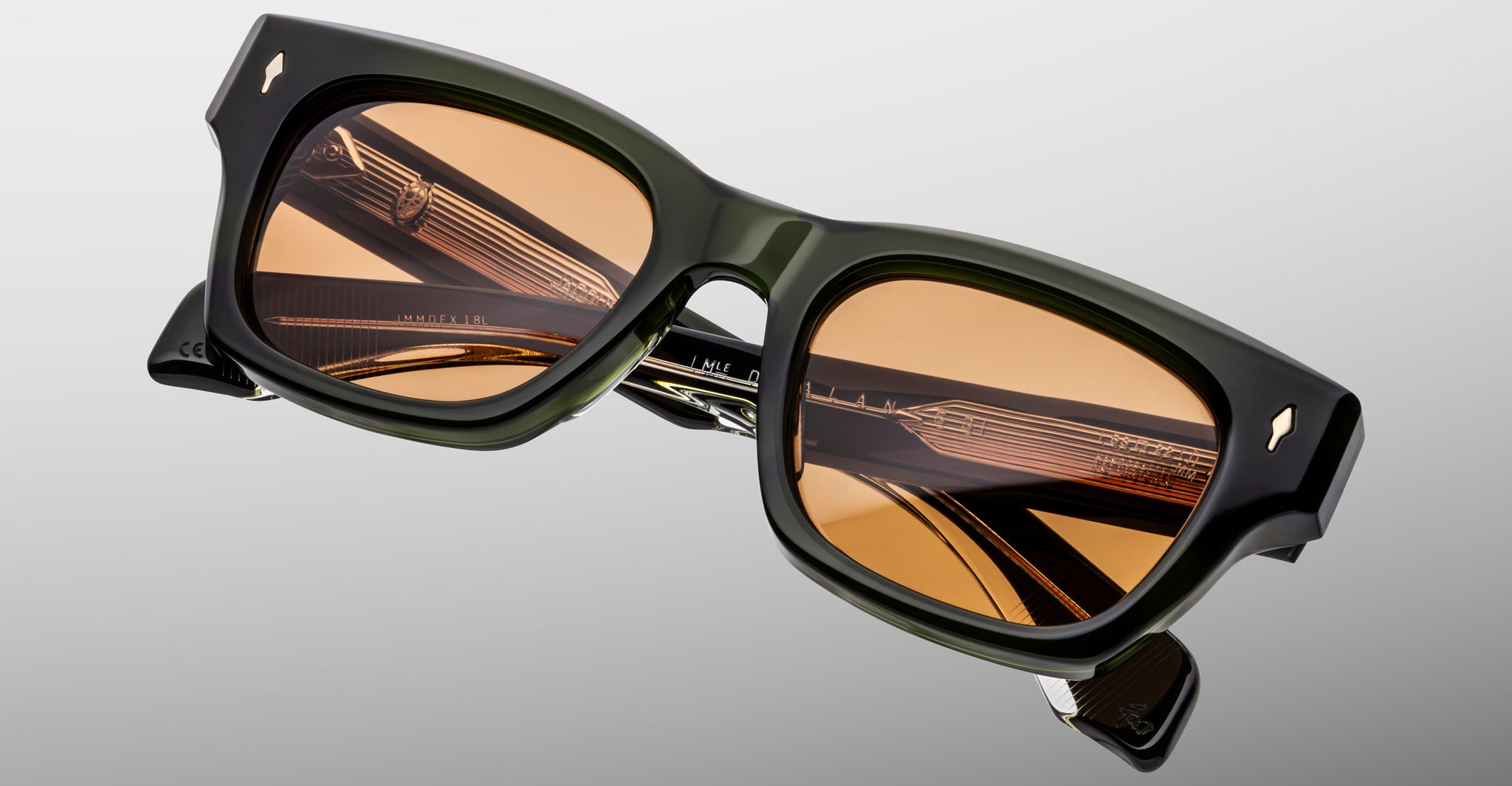 The Jacques Marie Mage Dealan 53 sunglasses feature chunky, rectangular dark green frames with amber-tinted lenses, gold detailing on the arms, and the brand’s logo near the hinges, shown against a light gray background.