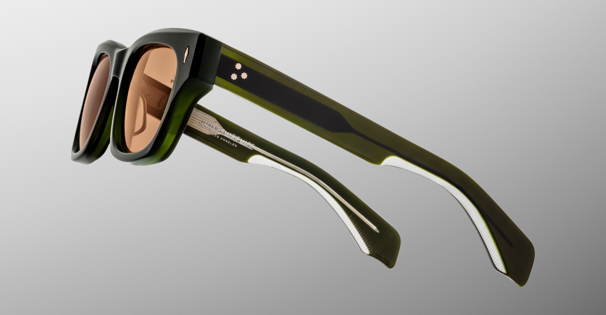 The Jacques Marie Mage Dealan 53 sunglasses feature thick, rectangular olive green frames, light brown lenses, and white arm detailing with two small metallic dots by the hinges, all set against a light gray background.