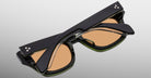 The Jacques Marie Mage Dealan 53, a pair of black sunglasses with thick frames and peach-tinted lenses, is folded and viewed from above on a light gray background, showing three small dots on the temples near the hinges.