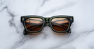 A pair of Jacques Marie Mage Dealan 53 dark square sunglasses with brown lenses rests folded on a white marble surface, showing reflections in the lenses.