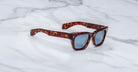 The Jacques Marie Mage Dealan 53 sunglasses, featuring thick tortoiseshell-patterned frames and dark lenses, are placed on a white marble surface with gray veining.