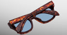 The Jacques Marie Mage Dealan 53 sunglasses, featuring a tortoiseshell pattern and blue lenses, are shown folded against a neutral gradient background.