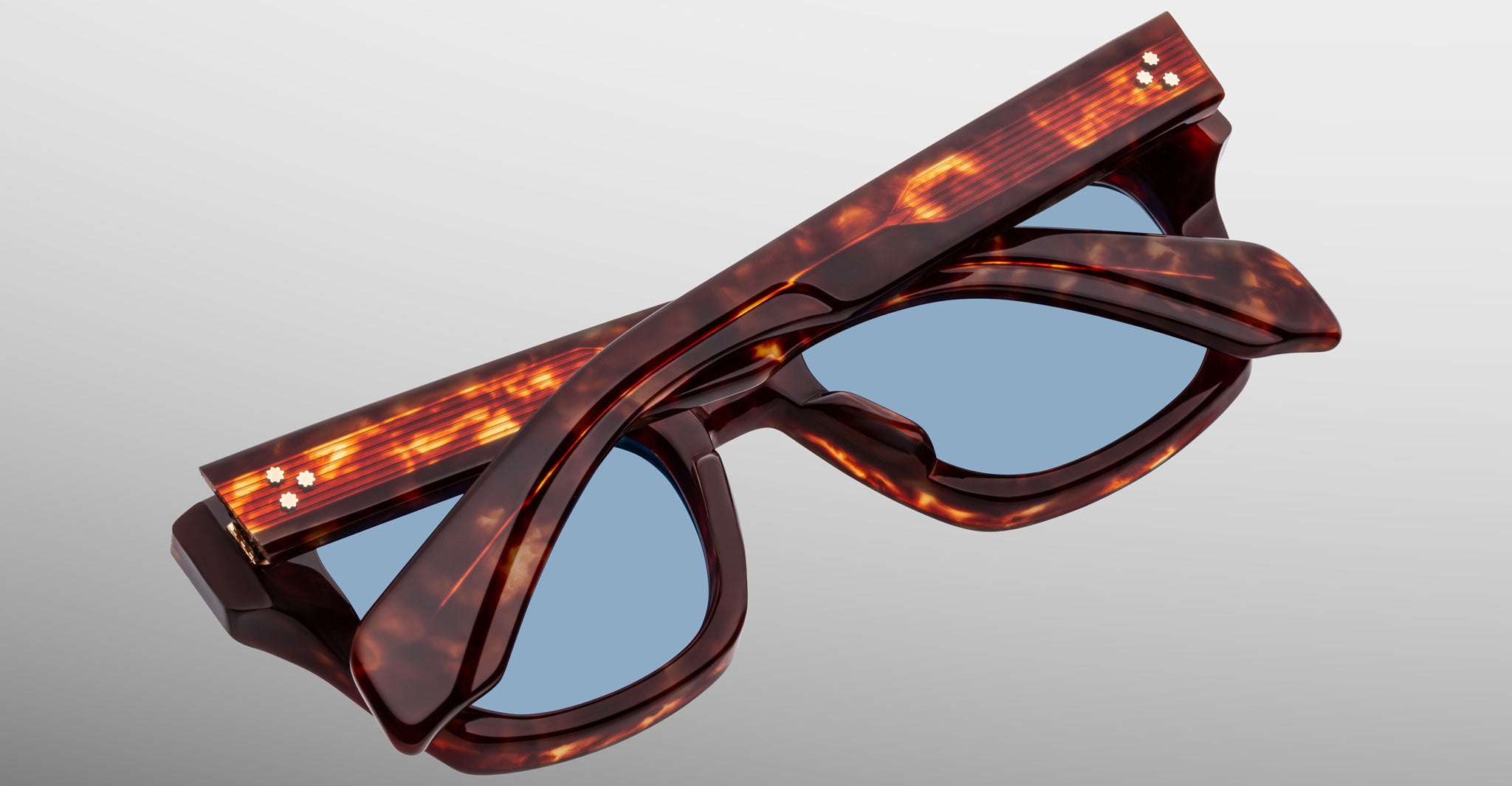 The Jacques Marie Mage Dealan 53 sunglasses, featuring a tortoiseshell pattern and blue lenses, are shown folded against a neutral gradient background.