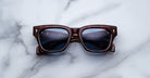 The Jacques Marie Mage Dealan 53, a pair of square dark brown tortoiseshell sunglasses with dark lenses, is displayed on a white marble surface.