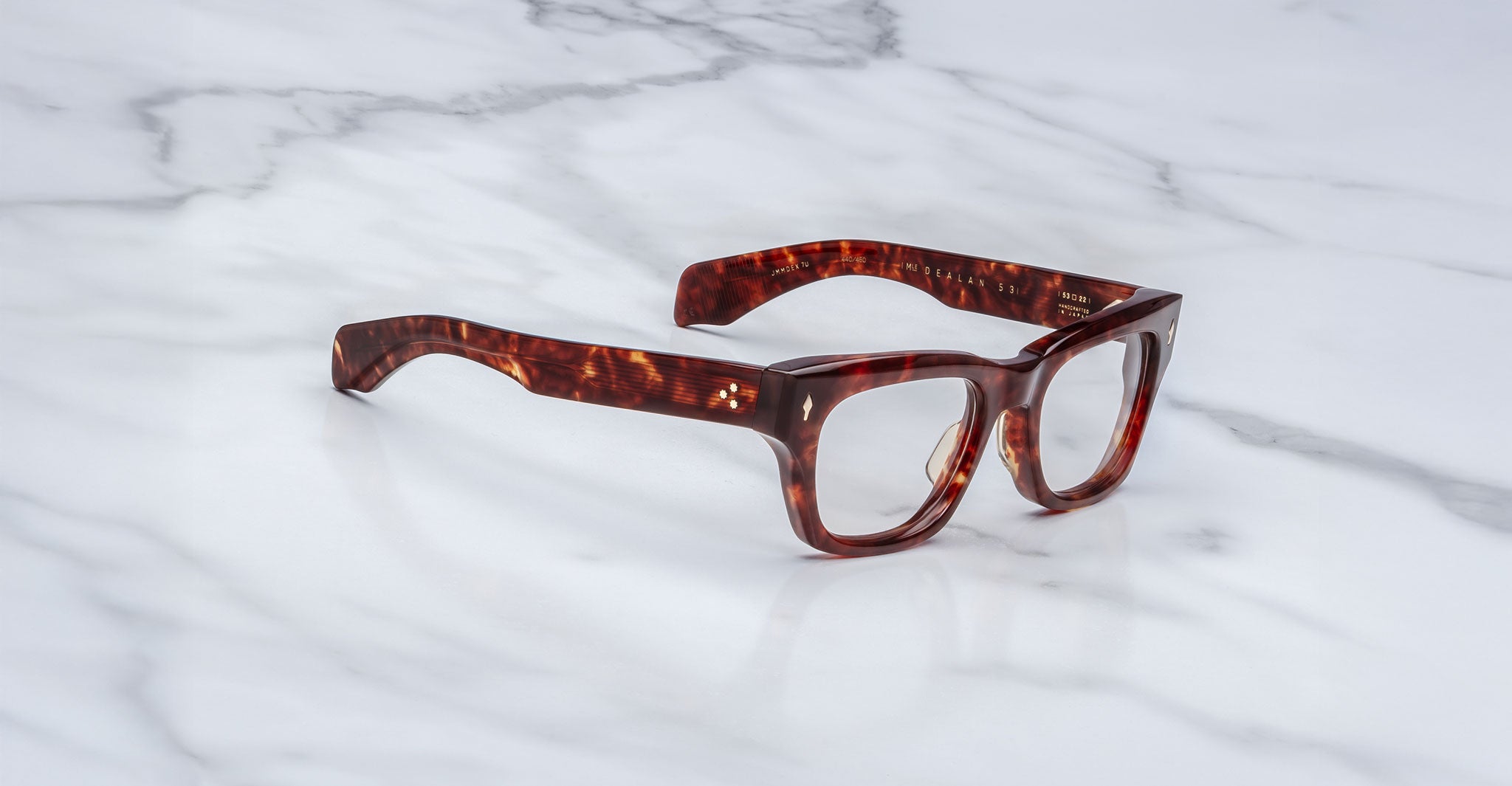 The Jacques Marie Mage Dealan 53 eyeglasses, featuring thick, rectangular tortoiseshell-patterned frames, are placed on a white marble surface.