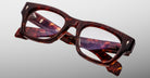 The Jacques Marie Mage Dealan 53 eyeglasses, featuring thick rectangular tortoiseshell frames with clear lenses, are displayed crossed on a light gray background, revealing gold inscriptions on the inner arms.