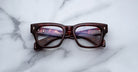 A pair of Jacques Marie Mage Dealan 53 rectangular eyeglasses with thick, dark tortoiseshell frames lies closed on a white marble surface.