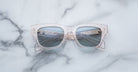 A pair of Jacques Marie Mage Dealan 53 clear, square-framed sunglasses with blue-tinted lenses rests on a white marble surface, the lenses reflecting a metal rail structure.