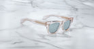 The Jacques Marie Mage Dealan 53 sunglasses, featuring clear frames and light blue-tinted lenses, rest on a white marble surface with grey veining. Gold accents highlight the arms near the hinges.