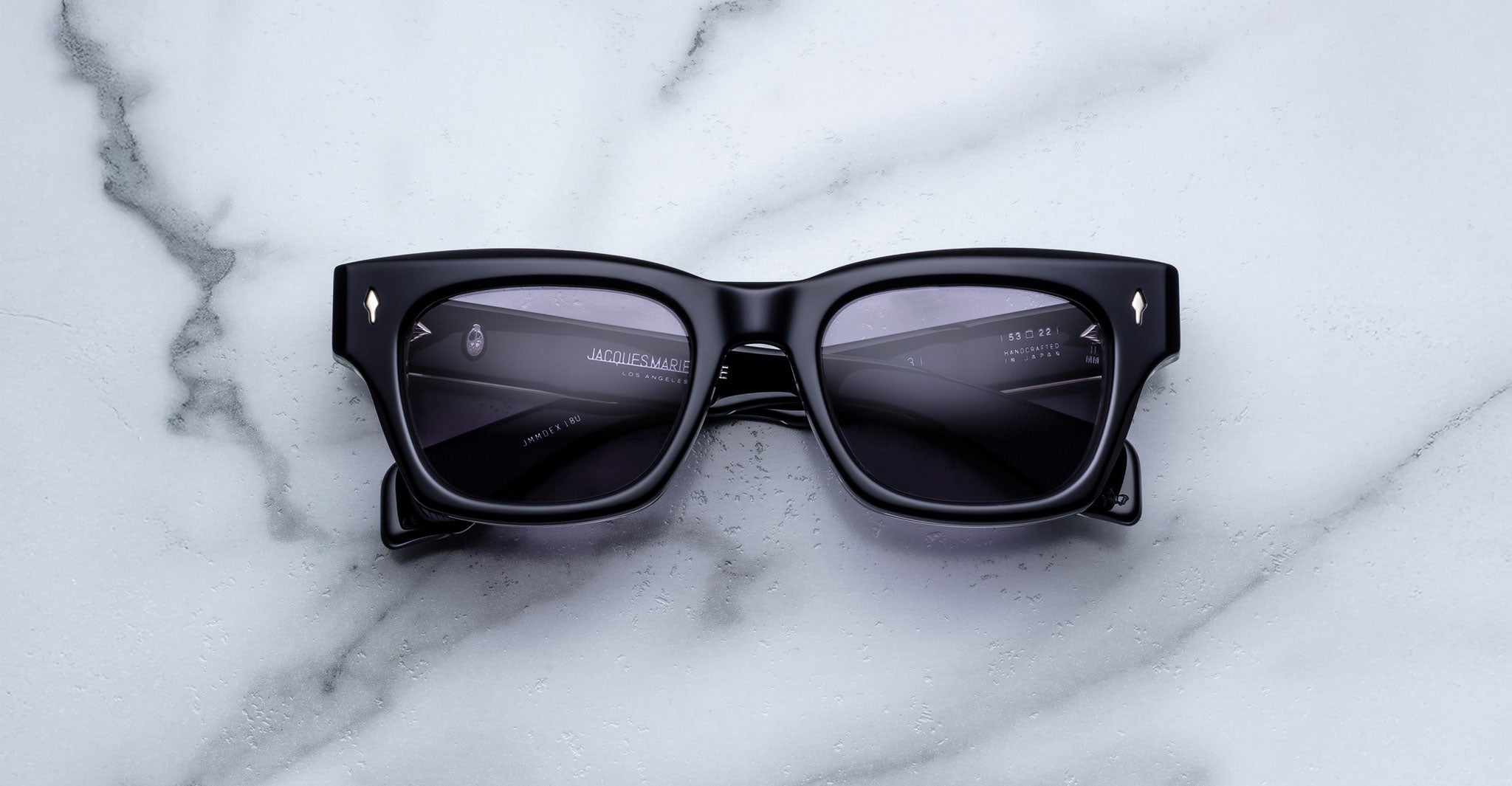 The Jacques Marie Mage Dealan 53 sunglasses, featuring black rectangular thick frames, rest on a white marble surface with reflections visible on the dark lenses.