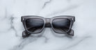 The Jacques Marie Mage Dealan 53 is a pair of black rectangular sunglasses with thick frames, shown face up on a white marble surface with folded arms.