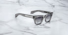 The Jacques Marie Mage Dealan 53 sunglasses, featuring gray thick frames and dark gradient lenses, rest on a white marble surface with gray veining.